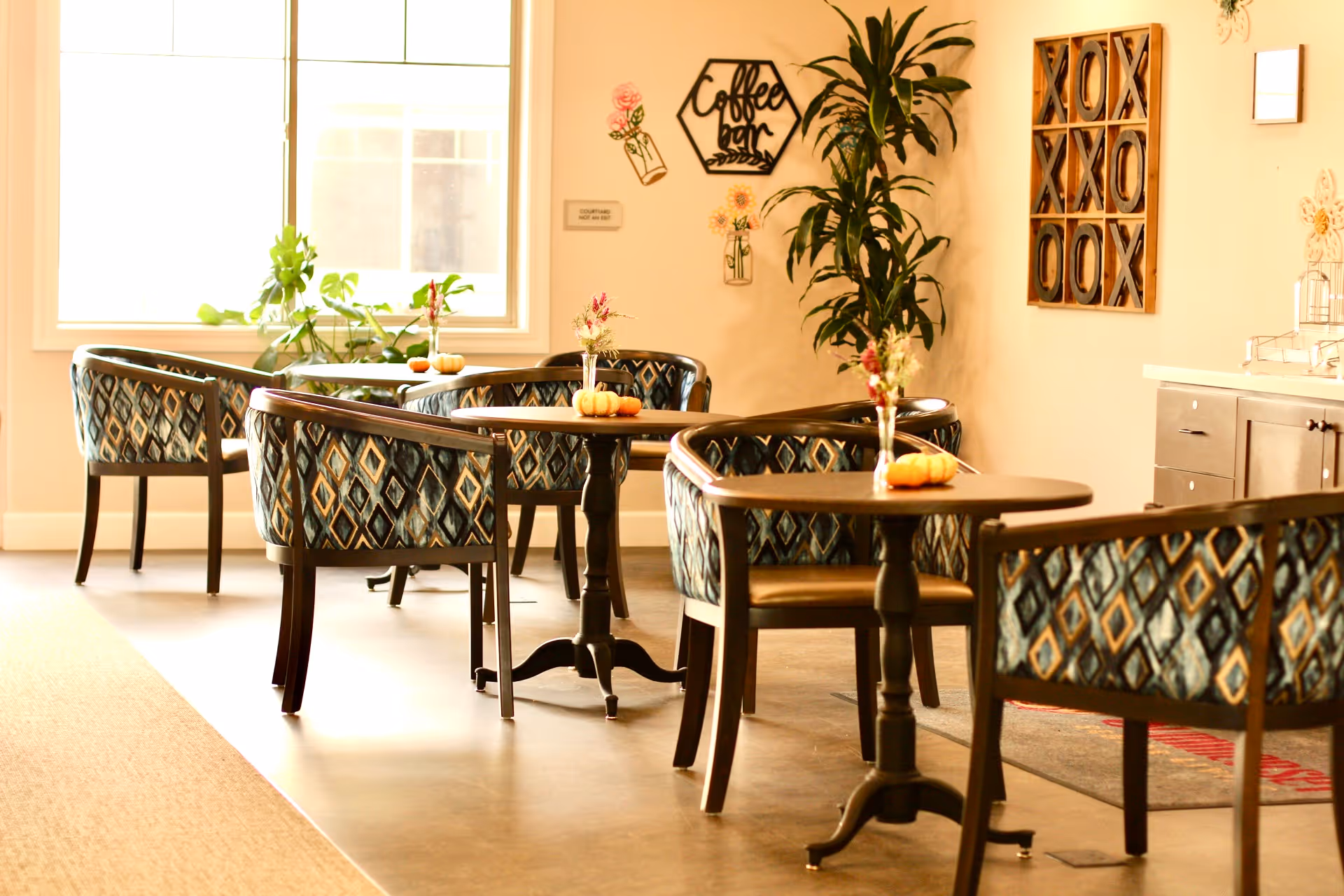 A cozy seating area with several small tables and patterned upholstered chairs arranged around them. Each table has a small decorative pumpkin and a vase with flowers. There is a large window letting in natural light, a tall green plant in the corner, and wall decorations including a sign that says 'Coffee bar' and a tic-tac-toe style wooden art piece.