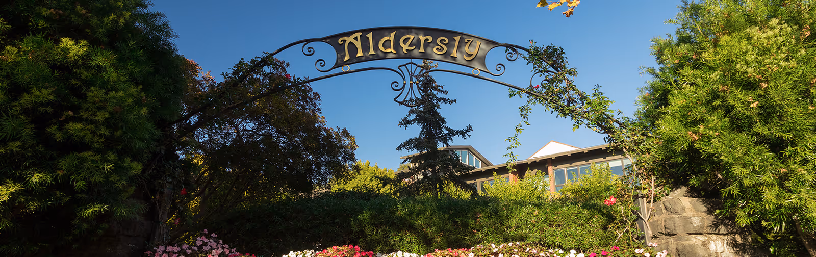A decorative metal archway with the name 'Aldersly' in gold letters, surrounded by lush green trees and colorful flowers under a clear blue sky. A building with large windows is visible in the background.