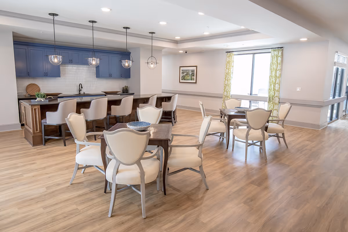 A bright and spacious dining area with wooden flooring, featuring two small square tables each surrounded by four cushioned chairs. In the background, there is a kitchen area with blue cabinets, a black countertop, a sink, and four bar stools. Three pendant lights hang above the kitchen counter. Large windows with green patterned curtains allow natural light into the room.