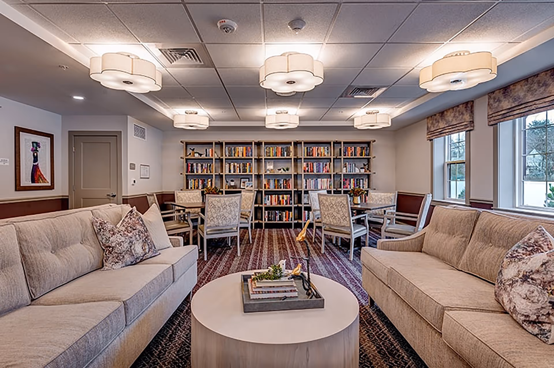 A cozy living room area with two beige sofas facing each other, a round white coffee table with decorative items in the center, and a bookshelf filled with books against the back wall. There are several tables with chairs arranged near the bookshelf, and large windows with patterned valances letting in natural light. The ceiling has multiple flower-shaped light fixtures.