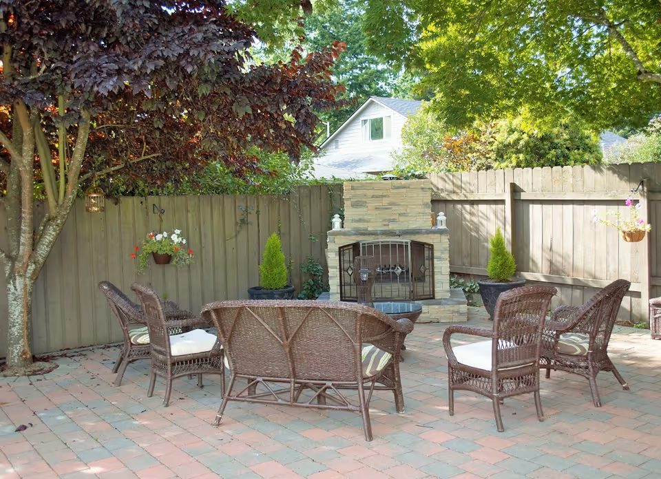 Outdoor patio area with wicker chairs and a loveseat arranged around a stone fireplace. The patio is paved with bricks and surrounded by a wooden fence with hanging flower pots and small potted evergreen plants. Trees provide shade over the seating area.