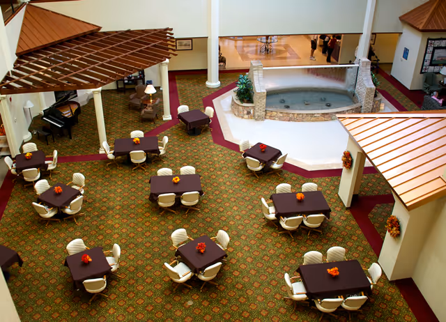 A spacious indoor dining area with multiple square tables covered with dark tablecloths, each surrounded by white chairs. The floor is carpeted with a patterned design, and there is a central water feature with a stone border. The area includes decorative elements such as a piano, lamps, and autumn-themed floral centerpieces on the tables. The ceiling has wooden pergola-style structures and white columns.