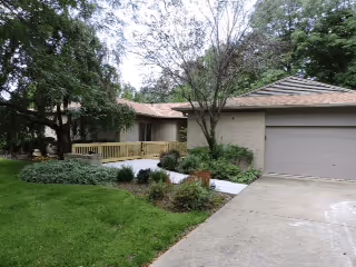 Exterior view of a single-story house with a garage on the right, a concrete driveway, and a front yard with green grass, bushes, and trees. The house has a light-colored exterior and a wooden ramp leading to the front door.