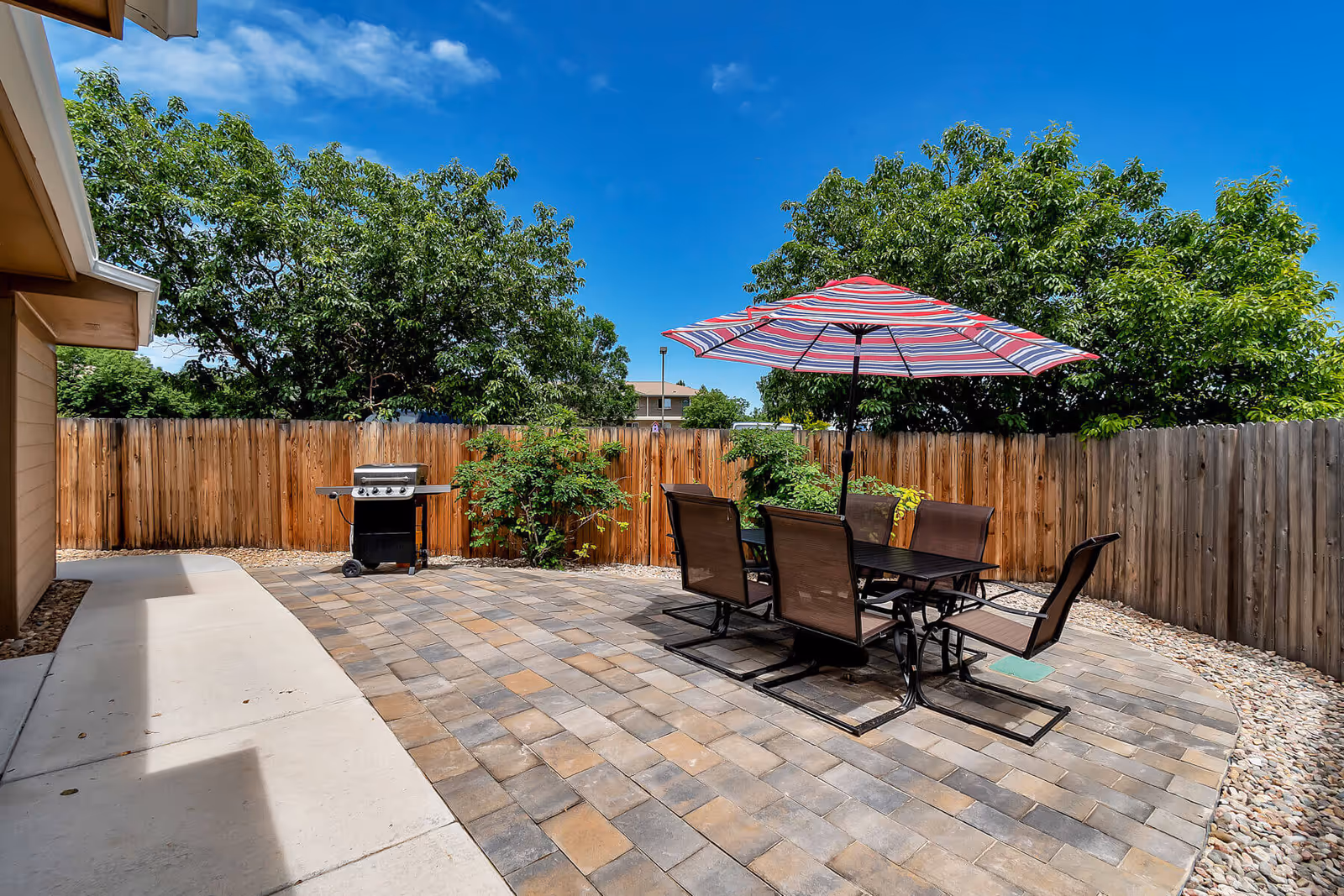 Outdoor patio area with a rectangular table and six chairs under a large striped umbrella. There is a barbecue grill against a wooden fence, with green trees and a clear blue sky in the background.