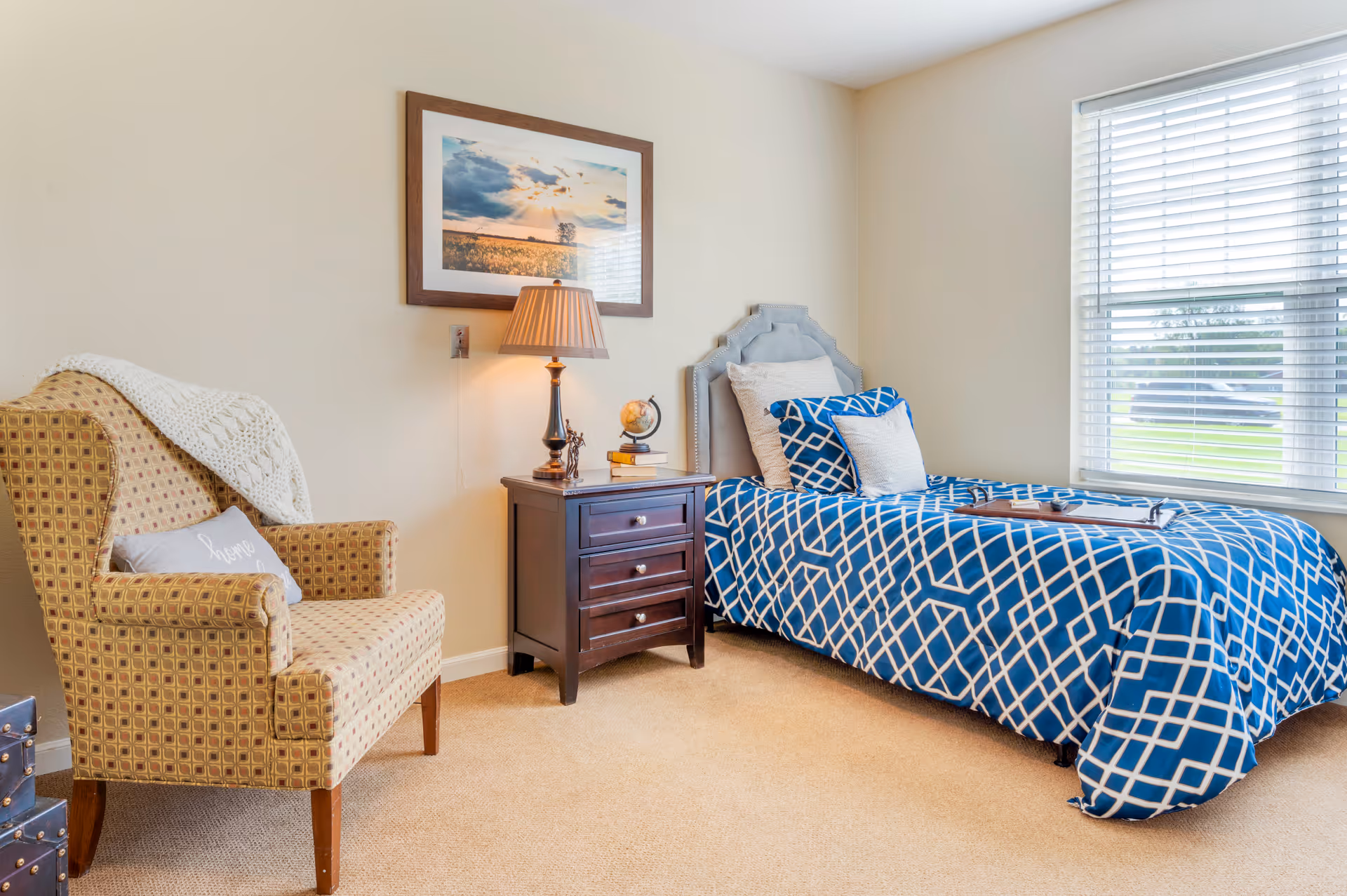 Bright bedroom with a twin bed dressed in blue geometric bedding, a dark wood nightstand with a lamp, and a patterned armchair near a window.
