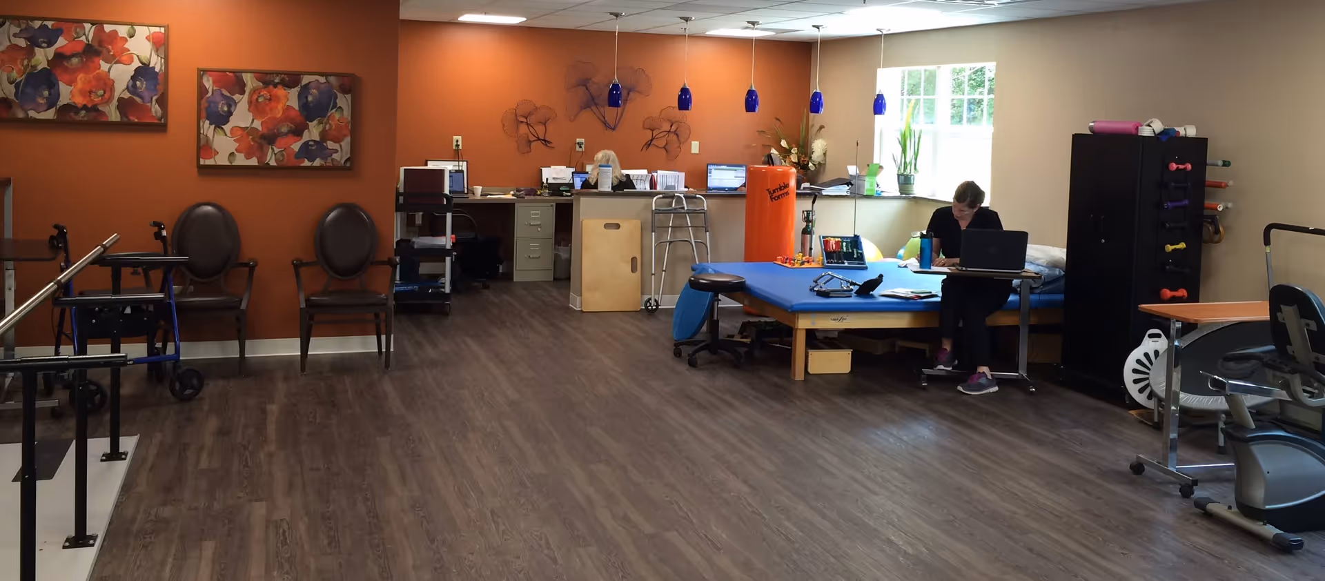 Rehabilitation therapy room with parallel bars, chairs, exercise equipment, a treatment table, and a staff member working at a desk.