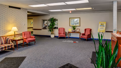 A spacious nursing home lobby area with blue carpet and beige wallpaper. The room features two red armchairs and a wooden table with a plant and framed picture on it. There is a striped sofa with a side table and lamp on the left side. A large potted plant is near the back wall, and another plant is in the foreground on the right. The ceiling has fluorescent lighting, and there is a sign on an easel near the back wall.