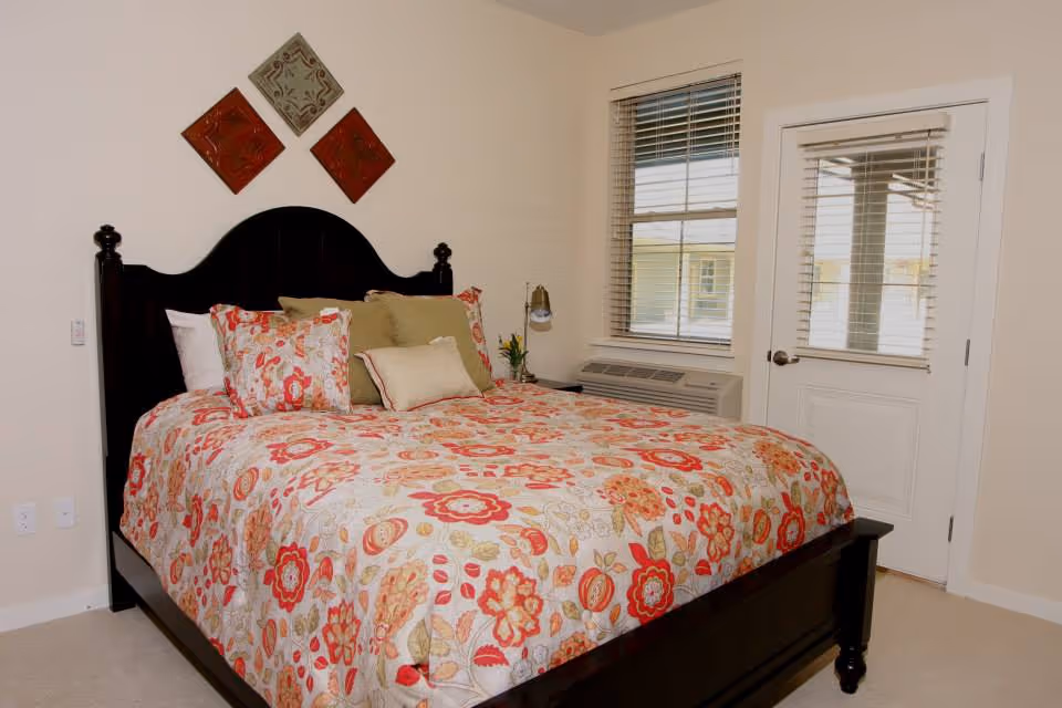 A bedroom with a dark wooden bed frame and a colorful floral bedspread in shades of red, orange, and beige. The bed has multiple pillows in coordinating colors. On the wall above the bed are three decorative square wall hangings arranged in a diamond pattern. To the right, there is a window with blinds and a door with a glass panel also covered by blinds. Below the window is an air conditioning unit.