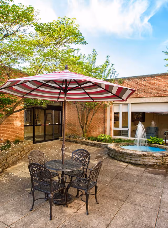 Outdoor patio area with a round metal table and four matching chairs under a large red and white striped umbrella. There is a small stone fountain with water jets on the right side, surrounded by a low stone wall. The patio is paved with square tiles and bordered by brick walls and greenery, including trees and plants. A glass door entrance to a building is visible in the background under a partly cloudy sky.