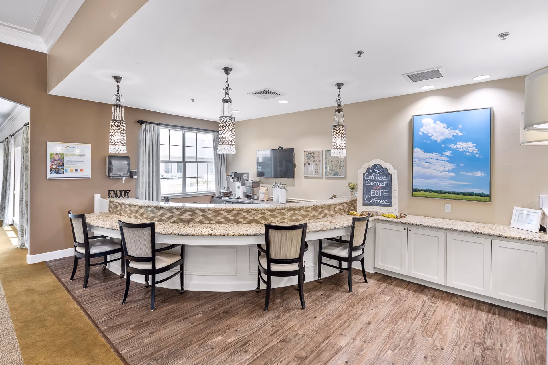 A bright and clean coffee corner area in a senior living facility with a curved granite countertop bar and four chairs. The space features hanging pendant lights, a window with curtains, a coffee machine, and a chalkboard sign that reads 'Coffee Corner EOTE Coffee.' There is a large painting of a blue sky with clouds on the wall and cabinets below the countertop.