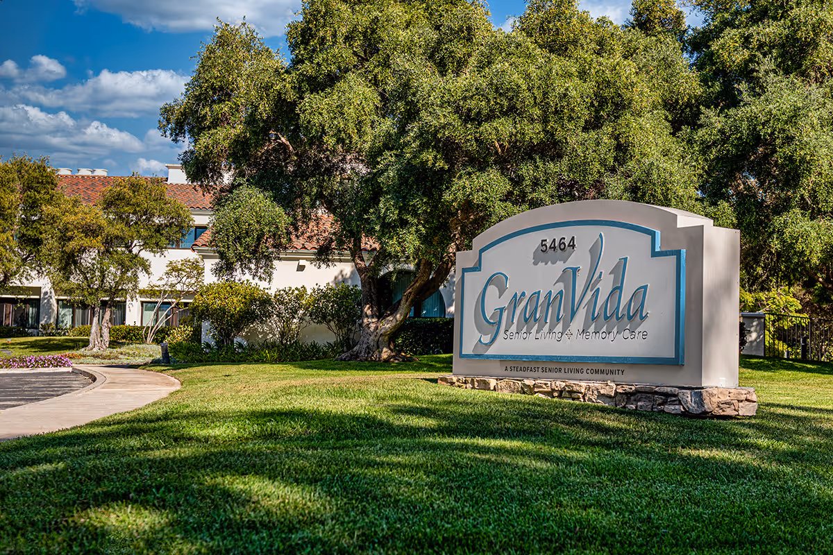 Outdoor view of the GranVida Senior Living and Memory Care facility sign with trees, grass, and part of the building with a red-tiled roof in the background under a partly cloudy sky.