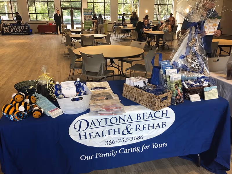 A promotional table for Daytona Beach Health & Rehab covered with a blue tablecloth displaying the facility's name, phone number, and slogan. The table holds various brochures, giveaways, and decorative items. In the background, there are round tables and chairs in a large, well-lit room with people seated and standing near other promotional booths.