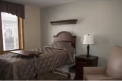 Private resident room with an adjustable bed, overbed table, bedside table with lamp, armchair, and a window with valance.