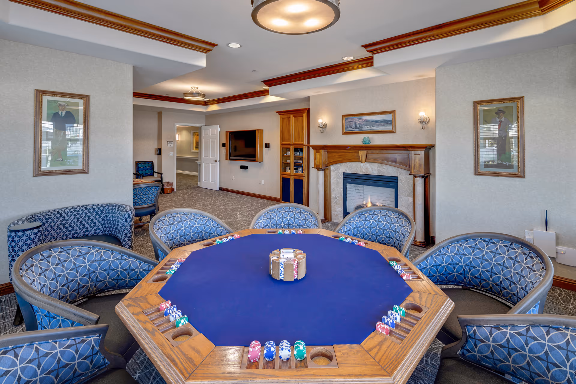 A cozy interior room featuring a large octagonal poker table with blue felt and poker chips arranged around it. Surrounding the table are cushioned chairs with a blue geometric pattern. The room has beige walls adorned with framed paintings, a fireplace with a wooden mantle, and a flat-screen TV mounted on the wall. The ceiling has recessed lighting and wooden crown molding.