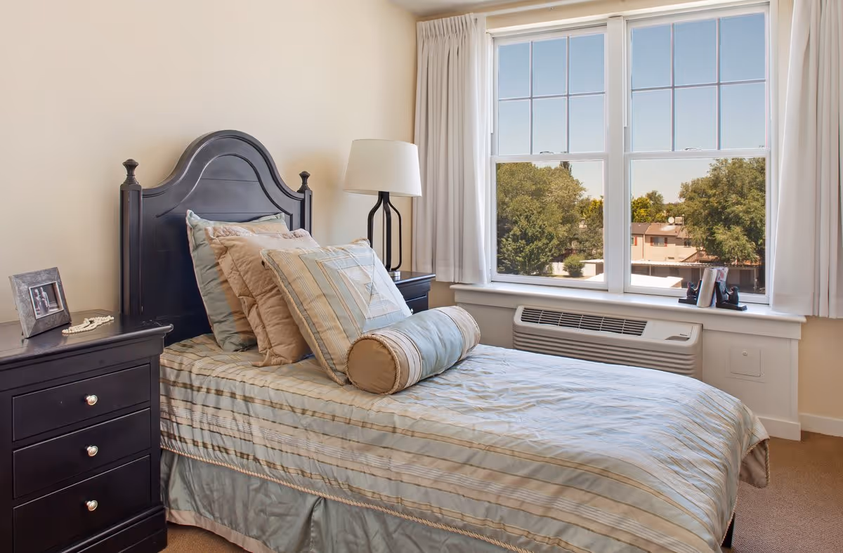 A neatly made single bed with multiple pillows and a bolster pillow in a bedroom. The bed has a dark wooden headboard and is next to a matching nightstand with a framed photo and a beaded necklace. A modern lamp is on another nightstand near a large window with white curtains, showing a view of trees and buildings outside.