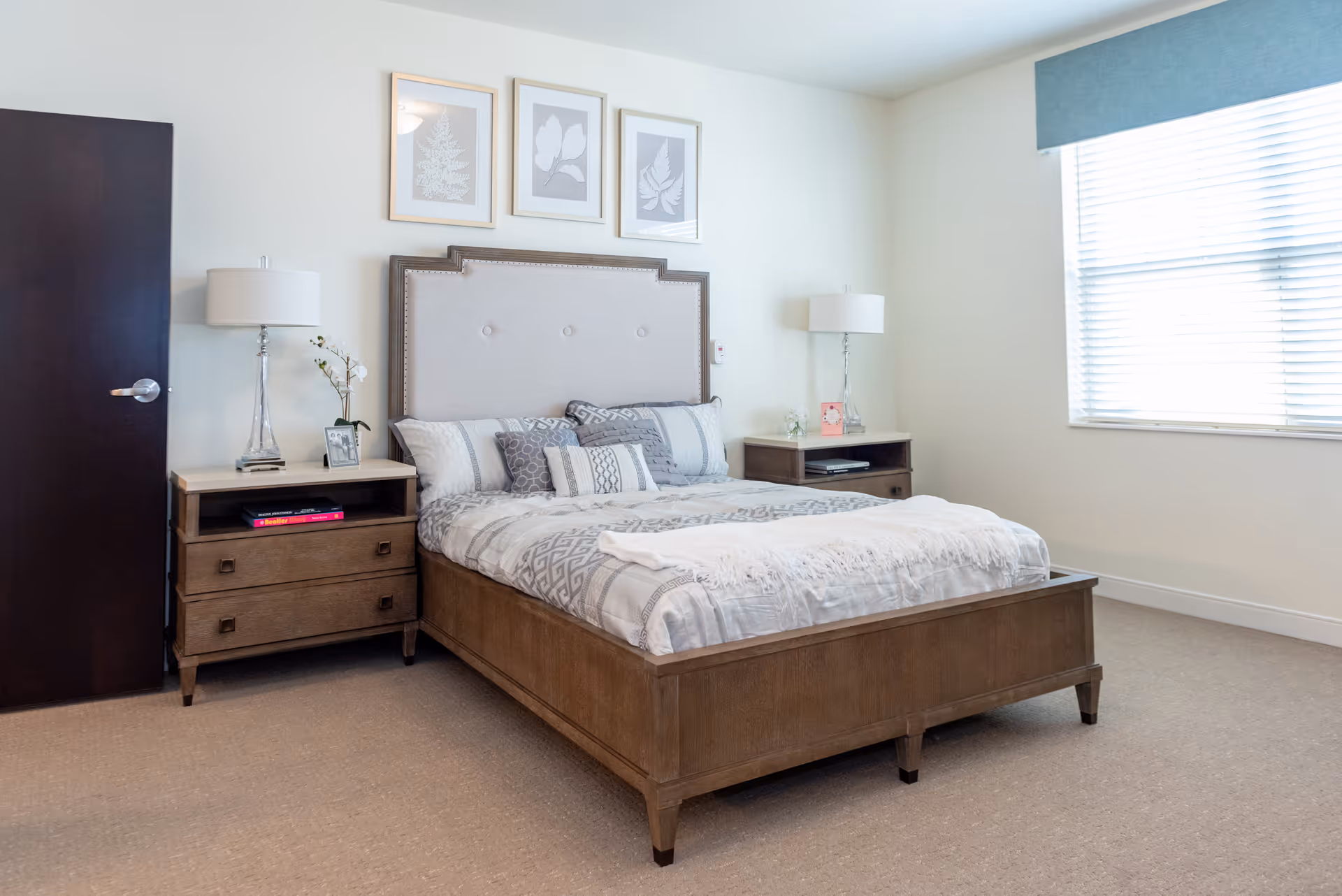 A neatly made bed with a wooden frame and upholstered headboard in a bright bedroom. There are two matching wooden nightstands on either side of the bed, each with a lamp and decorative items. Three framed botanical prints hang on the wall above the bed. A window with blinds and a blue valance lets in natural light.