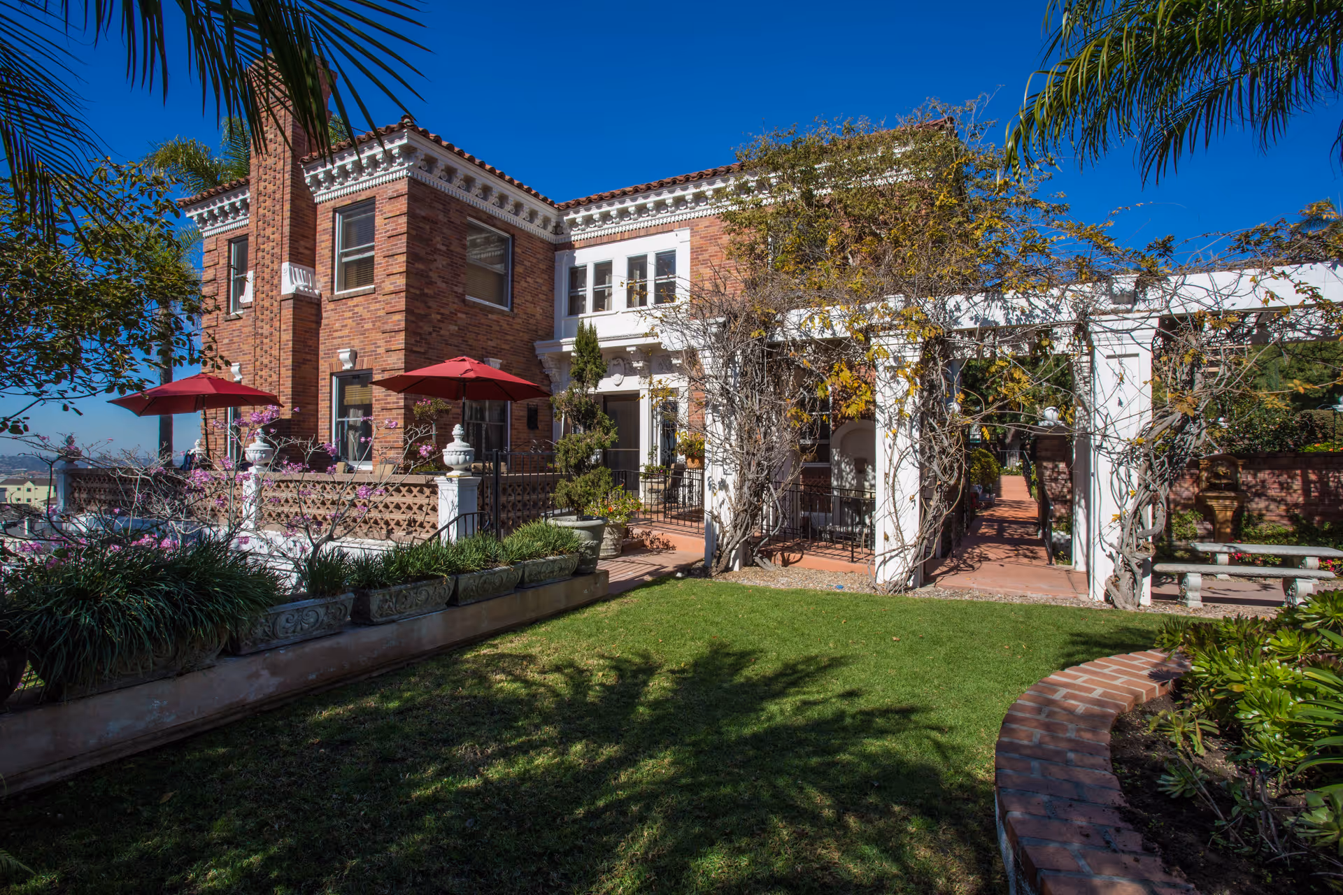 Brick two-story building with a tiled roof, outdoor patio with red umbrellas, vine-covered pergola, and a landscaped lawn.
