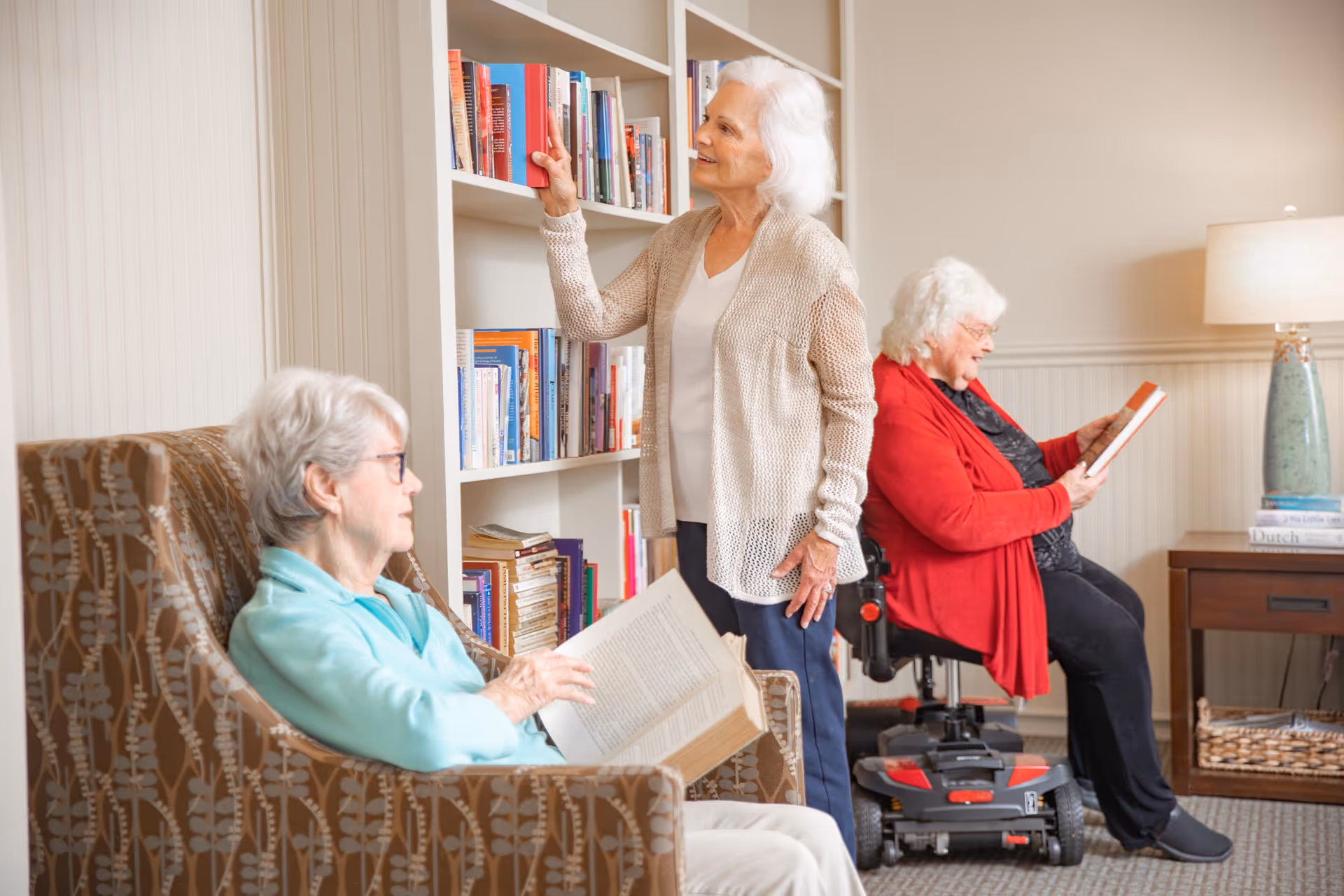 Three elderly women in a cozy room with bookshelves. One woman is seated in a patterned armchair reading a book, another woman is standing and reaching for a book on the shelf, and the third woman is sitting in a mobility scooter reading a book. The room has warm lighting and a side table with a lamp and stacked books.