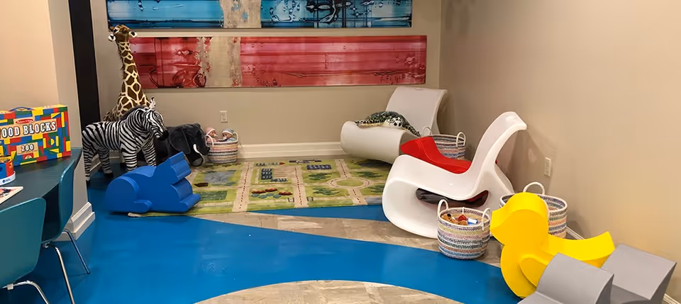A colorful playroom area with children's toys including a giraffe, zebra, elephant, and yellow duck, small chairs, baskets with toys, and a play mat with roads and buildings. The floor has blue and beige patterns, and there are abstract red and blue paintings on the wall.