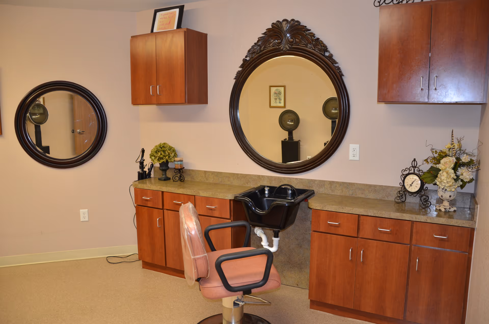 A salon station with two round mirrors, a shampoo sink, a styling chair, and wooden cabinets along a beige wall.