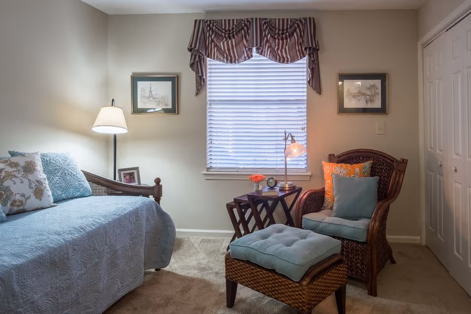 Cozy bedroom with a daybed on the left, a wicker armchair and ottoman on the right, a window with blinds and a small side table between them.