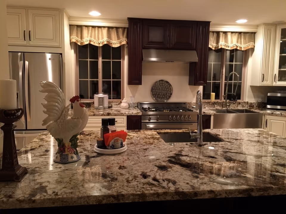 A modern kitchen with granite countertops, a stainless steel refrigerator, stove, and double sink. The kitchen island features a decorative ceramic rooster, a candle holder, and a small tray with condiments. There are two windows with valance curtains above the countertops.