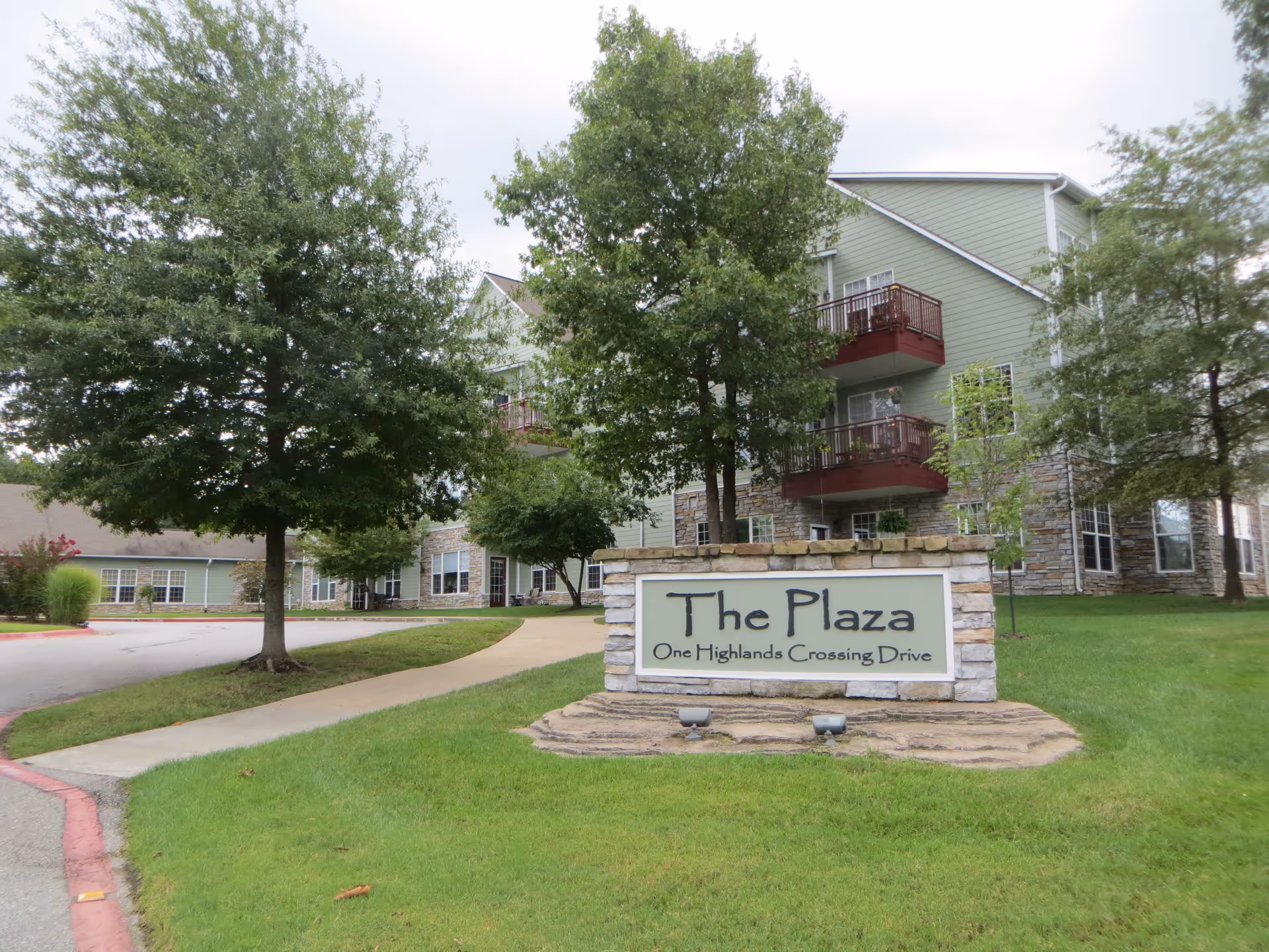 Exterior view of The Plaza at One Highlands Crossing Drive, showing a stone sign with the facility name in front of a green building with balconies and surrounded by trees and grass.