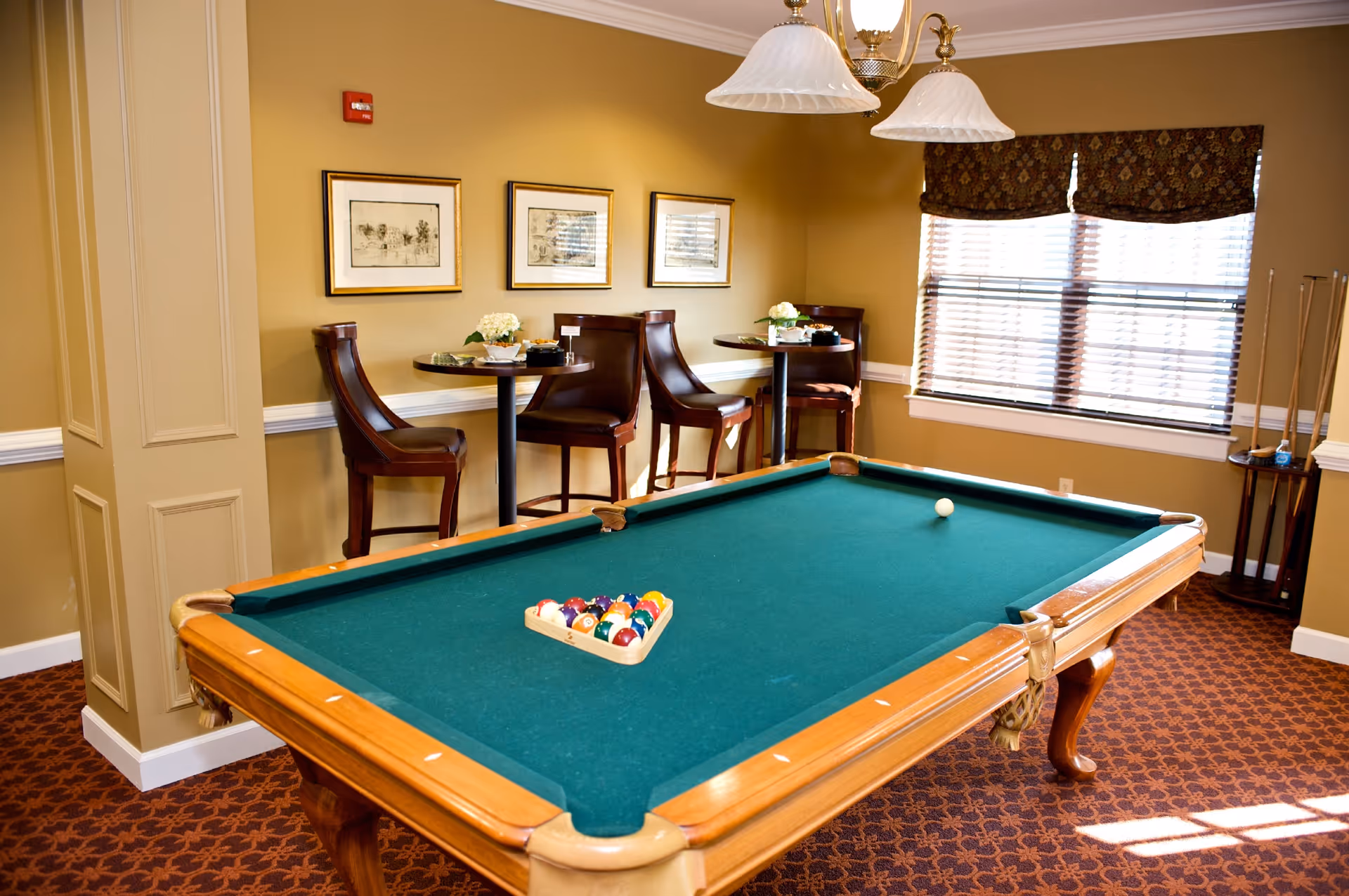 A well-lit room with a green felt pool table in the center, a rack of billiard balls arranged on the table, and a cue ball placed separately. Behind the pool table, there are two small round tables each with two high-back chairs. The walls are painted a warm beige color and decorated with three framed pictures. A window with patterned valances lets in natural light, and a chandelier with two lampshades hangs above the pool table. A corner stand holds pool cues.