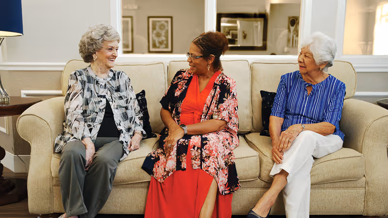 Three elderly women sitting on a beige couch in a living room, engaged in conversation and smiling. The room has neutral-colored walls, framed artwork, and a side table with a lamp.