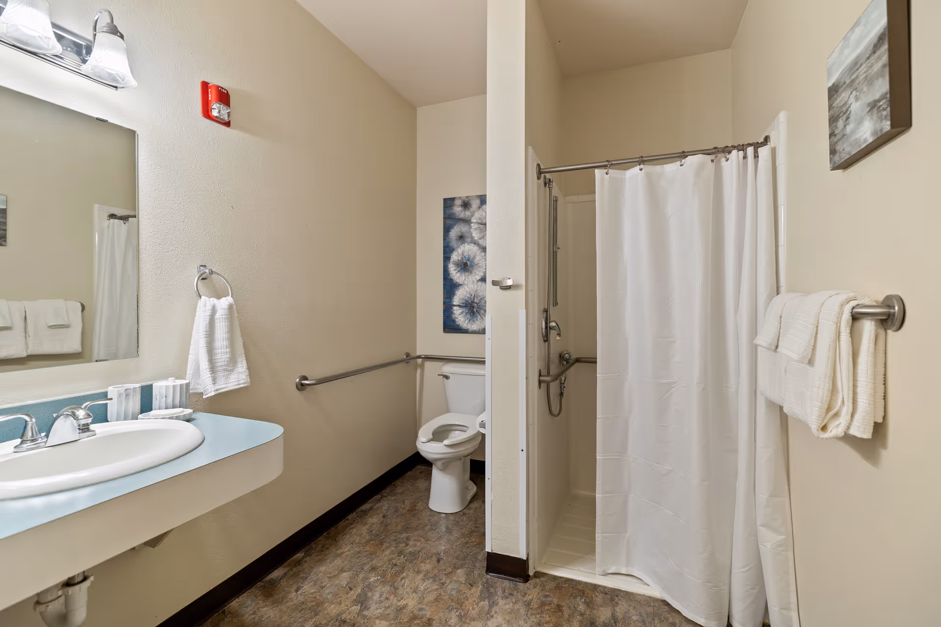 Accessible bathroom with a sink and mirror on the left, a toilet with grab bars in the center, and a shower with a white curtain on the right.