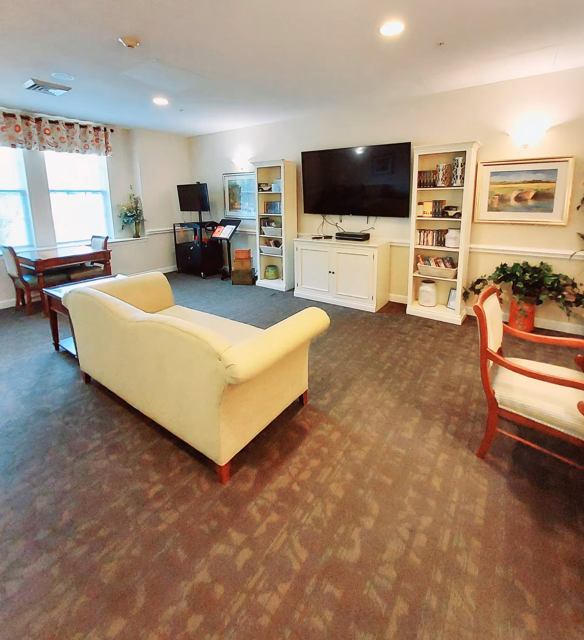 A cozy living room area with a cream-colored sofa facing a large flat-screen TV mounted on the wall. There are two white bookshelves flanking the TV, filled with books and decorative items. To the left, there is a small wooden table with chairs near two large windows with floral valances. The room has carpeted flooring and soft lighting, with a framed painting and a potted plant on the right side.