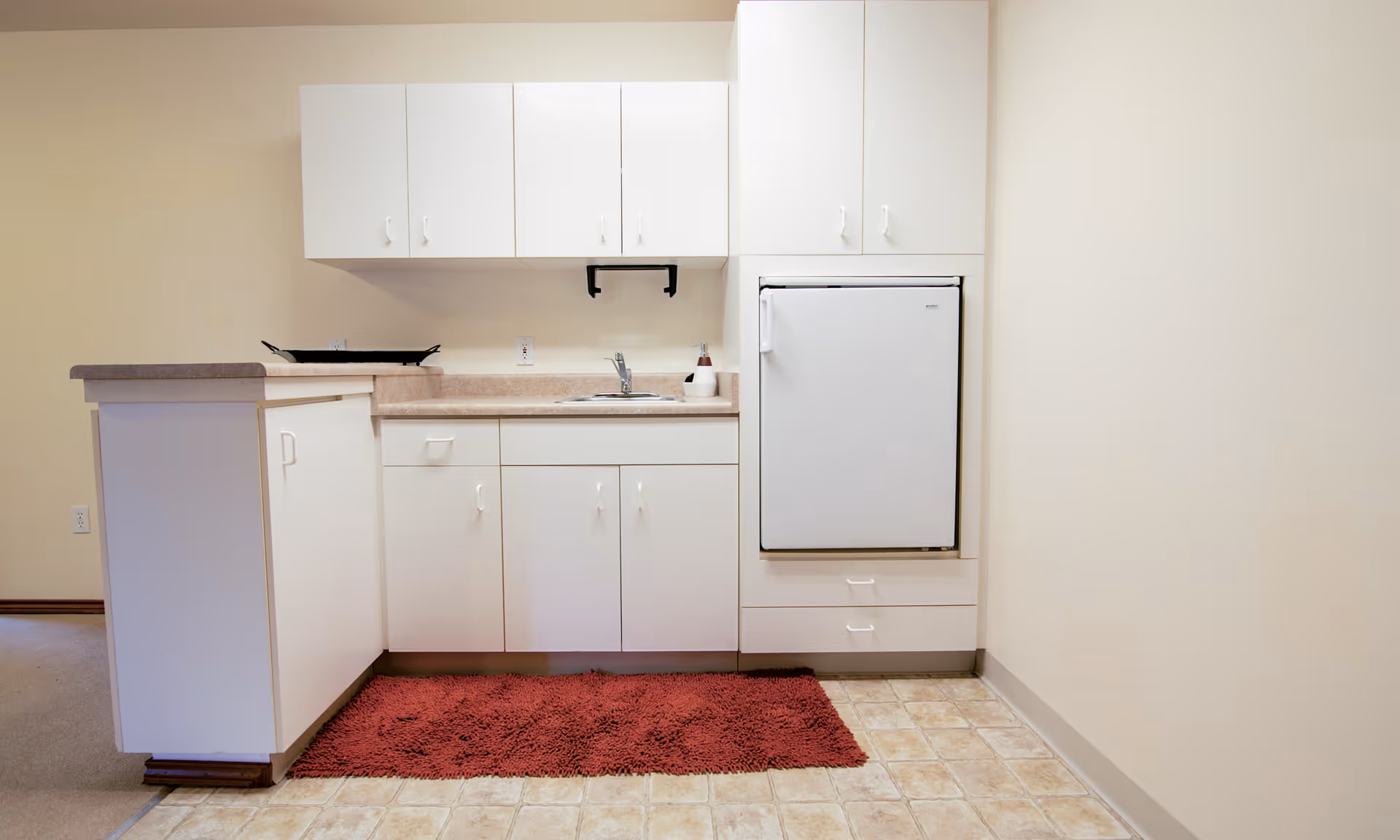 Small kitchenette with white cabinets, countertop, sink, mini refrigerator and a red rug on the floor.