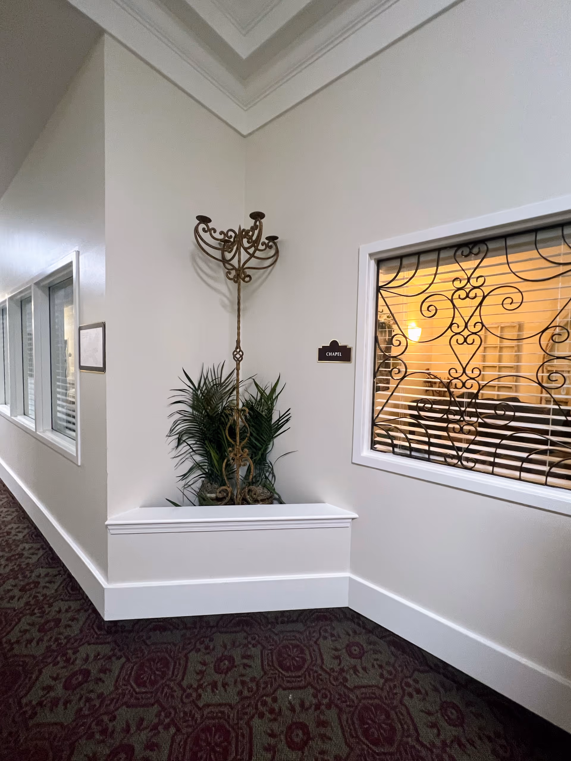 Interior corner of a hallway with white walls and patterned carpet. A decorative metal stand with candle holders is placed in a planter with green plants. A window with ornate metal grillwork looks into a warmly lit room labeled 'Chapel' on a small sign beside the window.