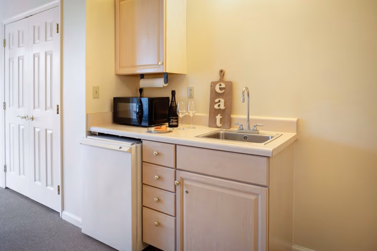 A small kitchenette area with light wood cabinets, a white countertop, a stainless steel sink with a faucet, a black microwave, a mini refrigerator, a bottle of wine, two wine glasses, a plate with food, and a wooden cutting board with the word 'eat' on it. There are white double doors to the left and beige walls.