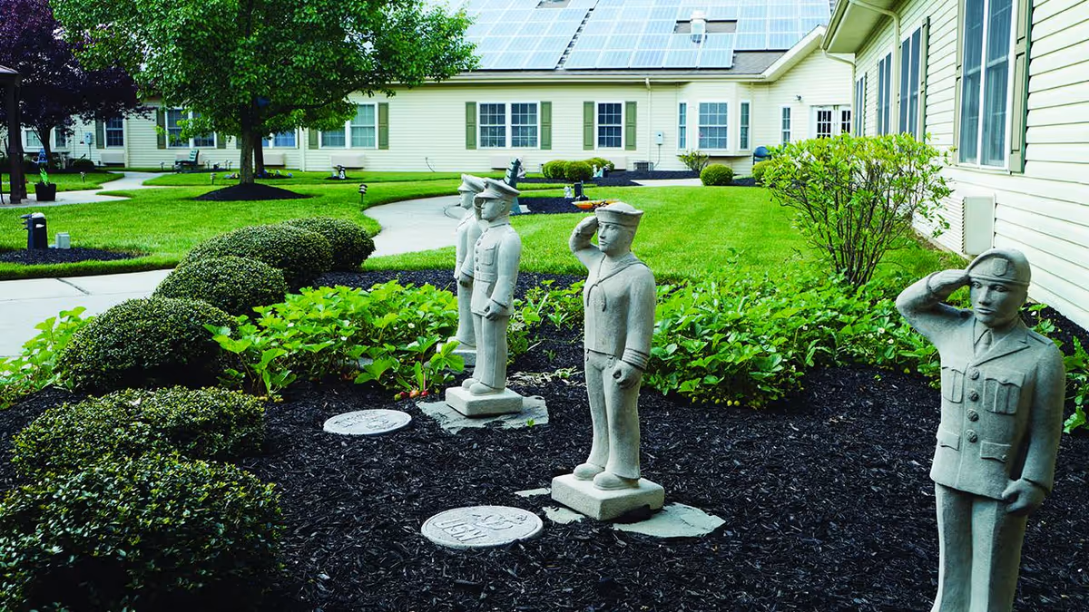Outdoor garden area at Traditions of Cross Keys featuring neatly trimmed bushes, green plants, and three stone statues of saluting military figures on a bed of dark mulch, with a light-colored building and paved walkways in the background.