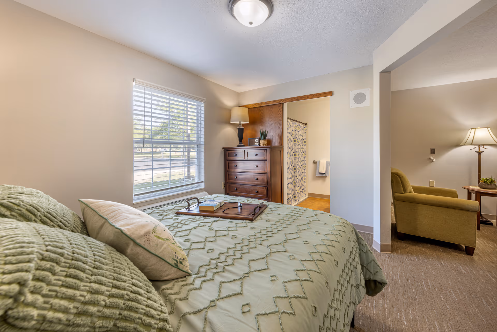 A bedroom with a bed covered in a green textured bedspread and pillows. A wooden dresser with a lamp and small plants sits against the wall near a window with blinds. An open doorway reveals a bathroom with a patterned shower curtain and towel hanging on a rack. To the right, a partial view of a living area with an armchair, side table, and lamp is visible.