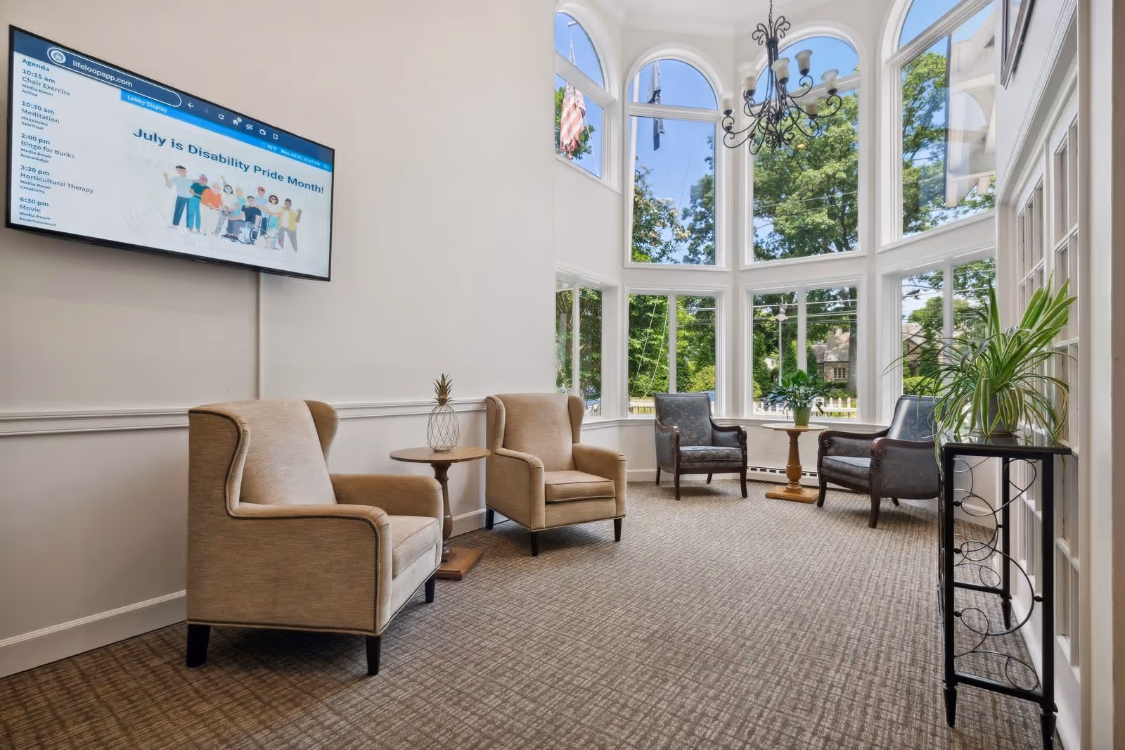 A bright and spacious sitting area with large windows letting in natural light and offering a view of green trees outside. The room features two beige armchairs with a small round wooden table between them, two dark-colored armchairs with another small round table between them near the windows, a chandelier hanging from the ceiling, and a flat-screen TV mounted on the wall displaying a message about Disability Pride Month. There is also a black metal plant stand with a green plant on it near the door.