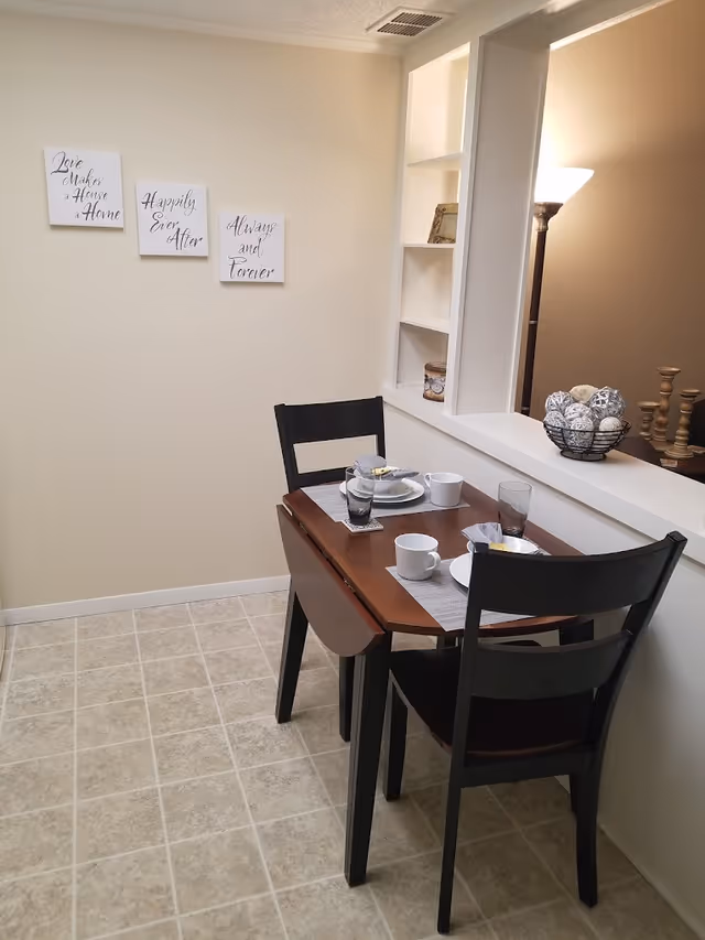 A small dining area with a wooden table set for two with plates, cups, and glasses. Two black chairs are placed at the table. On the wall, there are three decorative signs with the phrases 'Love makes a House a Home,' 'Happily Ever After,' and 'Always and Forever.' A built-in shelf with decorative items and a floor lamp are visible in the background.