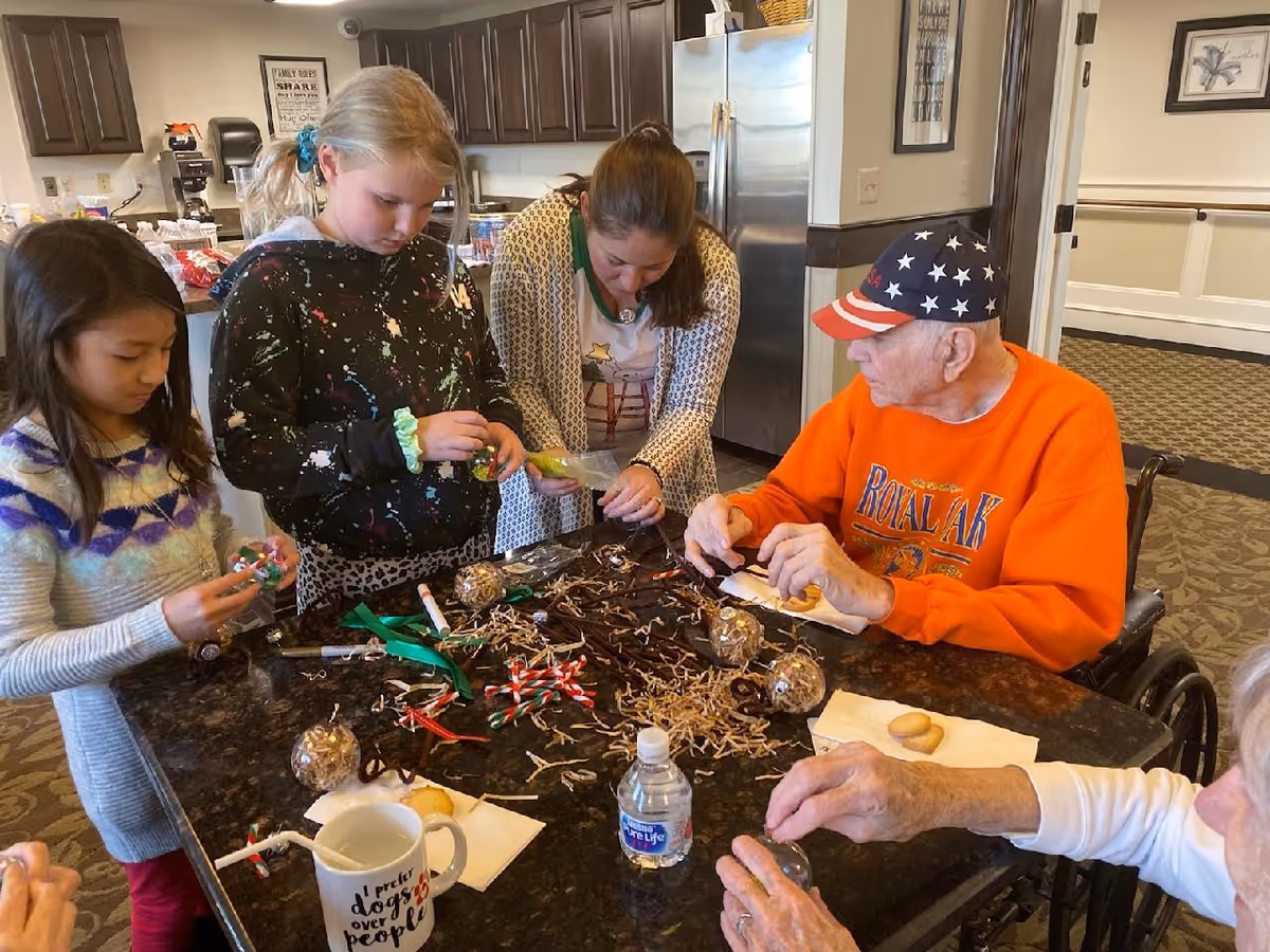 A group of children and elderly people sitting and standing around a table in a kitchen area, engaging in a craft activity with various materials like ribbons, pipe cleaners, and small ornaments. The elderly man in an orange sweatshirt and star-spangled cap is seated in a wheelchair. The kitchen has dark wooden cabinets and a stainless steel refrigerator in the background.