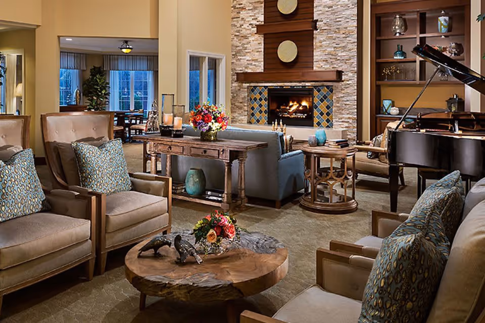 A cozy living room with beige armchairs and sofas adorned with patterned cushions, a wooden coffee table with decorative items and flowers, a stone fireplace with a fire burning, built-in shelves with decorative objects, and a grand piano on the right side. In the background, there is a dining area with large windows and a plant.