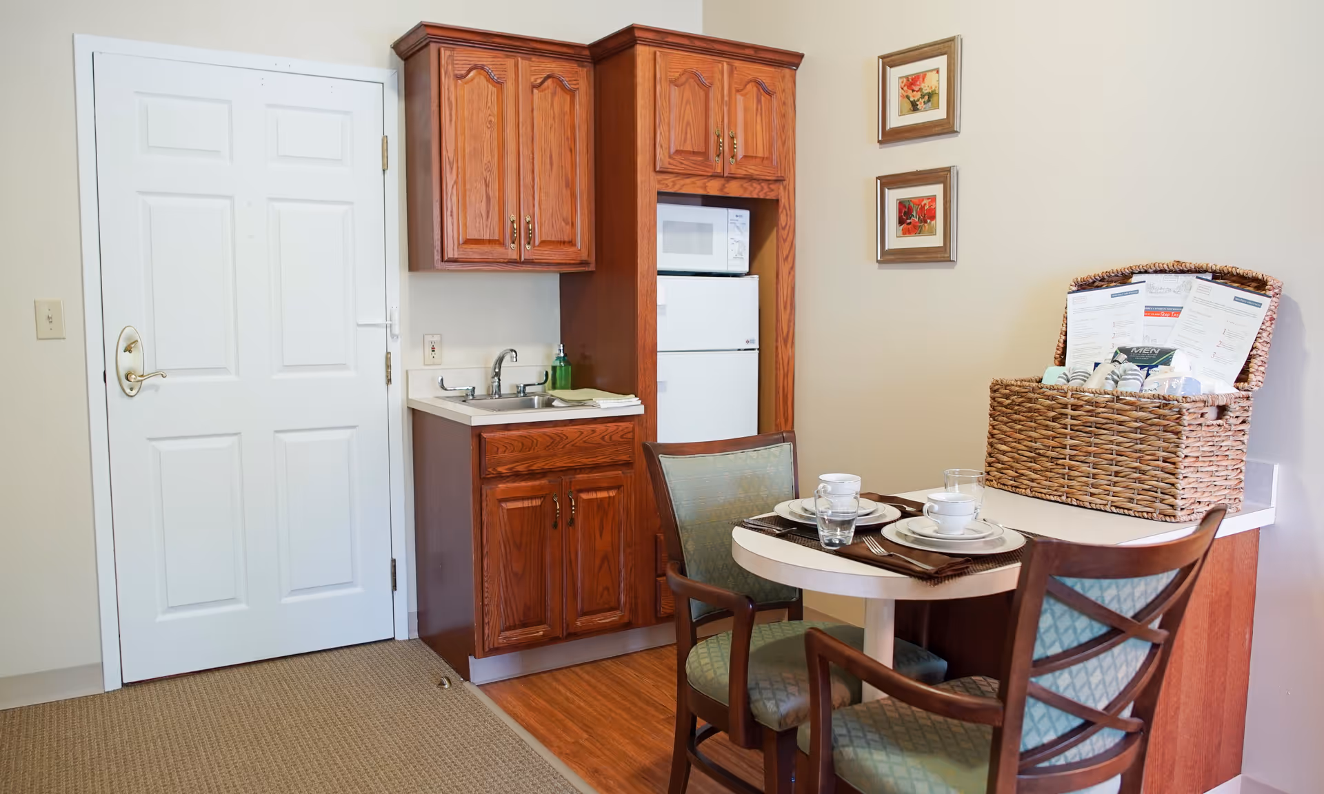 A small kitchenette area with wooden cabinets, a white microwave, and a white mini refrigerator. There is a sink with a green soap dispenser and a folded towel on the counter. Next to the kitchenette is a small round dining table set for two with plates, cups, and glasses. A wicker basket filled with various items and papers is placed on the counter extension. Two framed floral pictures hang on the beige wall above the basket. A white door is visible to the left of the kitchenette.