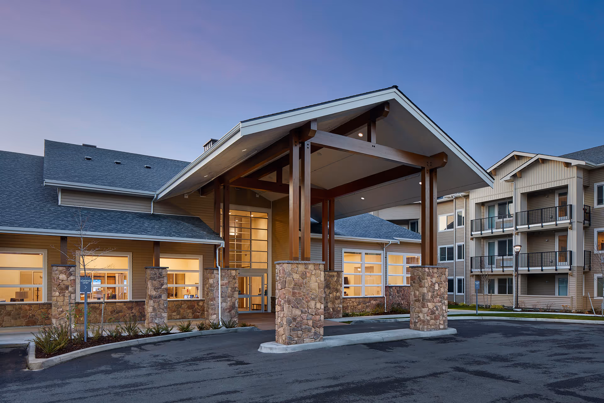 Front entrance of a senior living facility with a large covered porte-cochère supported by wooden beams and stone pillars and adjoining multi-story residential wings with balconies.