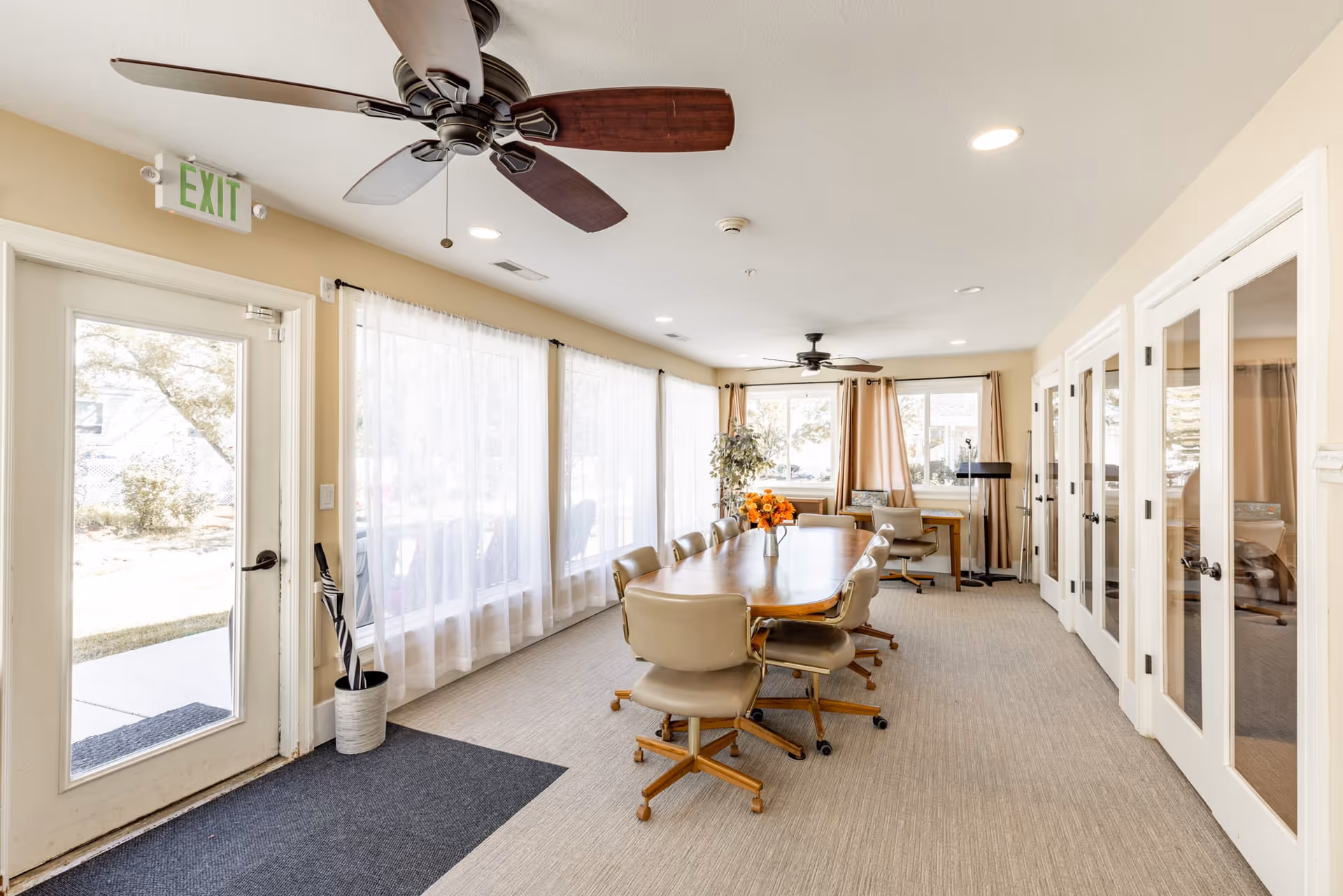 Bright communal dining/meeting room with a long wooden table surrounded by swivel chairs, large windows with sheer curtains, ceiling fans, and an exit door.