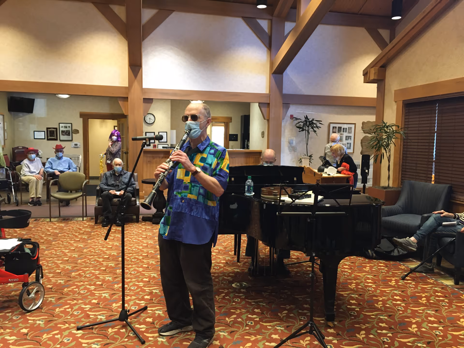 An elderly man wearing a colorful shirt, sunglasses, and a face mask plays a clarinet in a senior living facility common area. Behind him, another man plays a grand piano. Several elderly residents, some wearing face masks and festive hats, sit and watch the performance. The room has wooden beams, framed pictures on the walls, and patterned carpet.