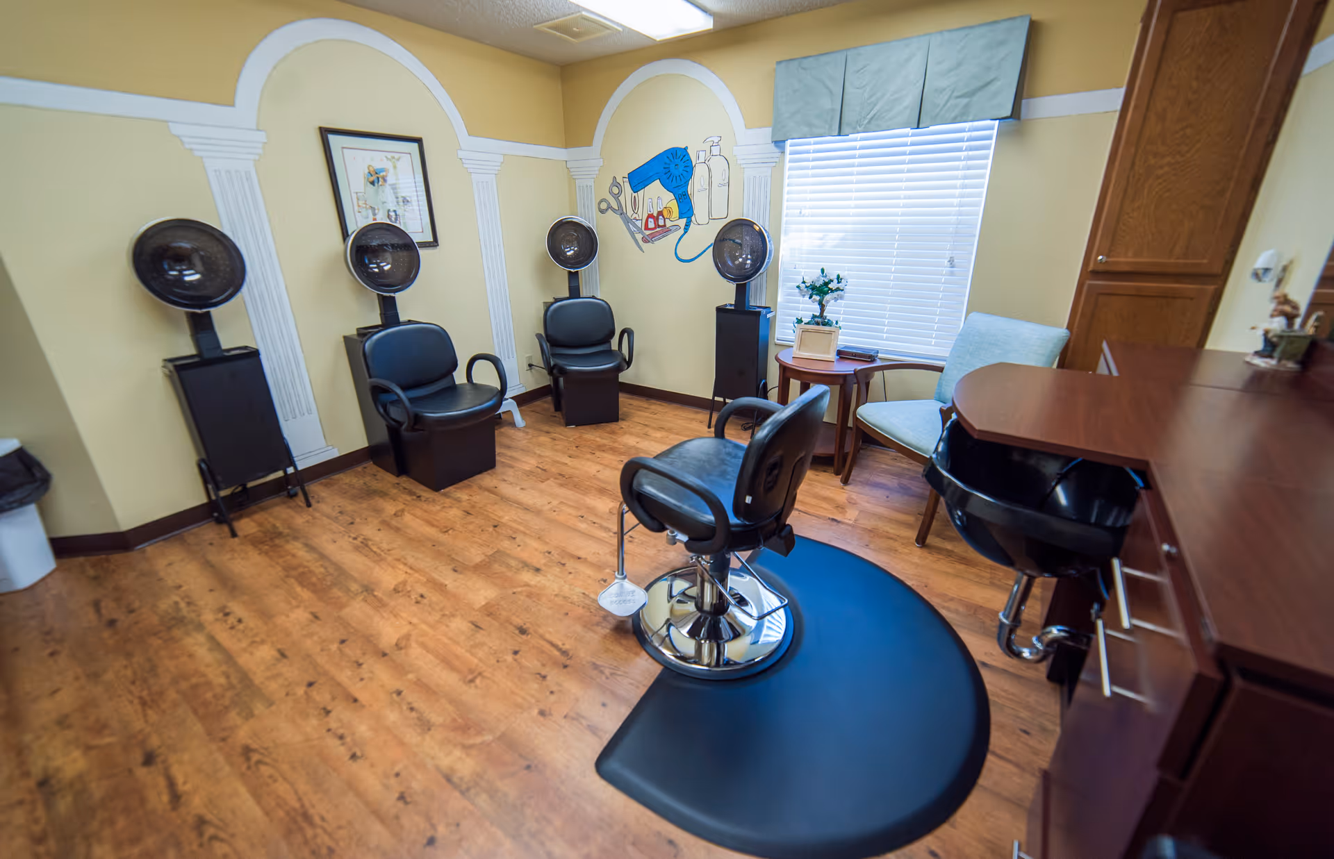 A bright salon room with multiple hooded hair dryers, styling chairs, a shampoo sink, and wood cabinetry.