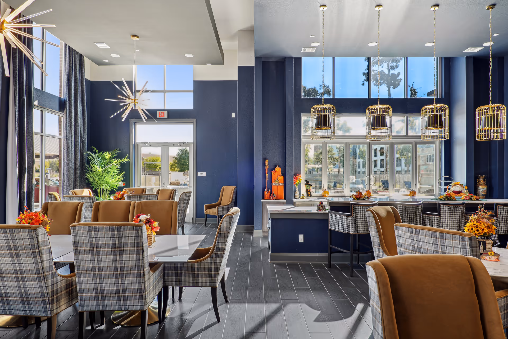 A bright and modern dining area with large windows letting in natural light. The room features multiple tables with plaid and brown upholstered chairs, decorative flower arrangements on the tables, and stylish pendant lights hanging from the ceiling. There is a bar area with high chairs and large windows behind it, and the walls are painted dark blue.