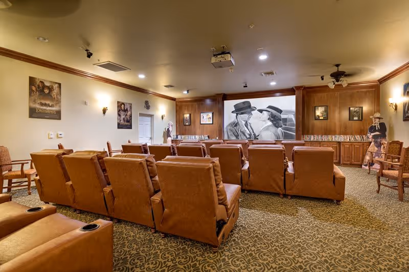 A cozy theater-style media room with rows of brown leather recliners facing a projector screen showing a black-and-white movie, wood-paneled walls and movie posters.
