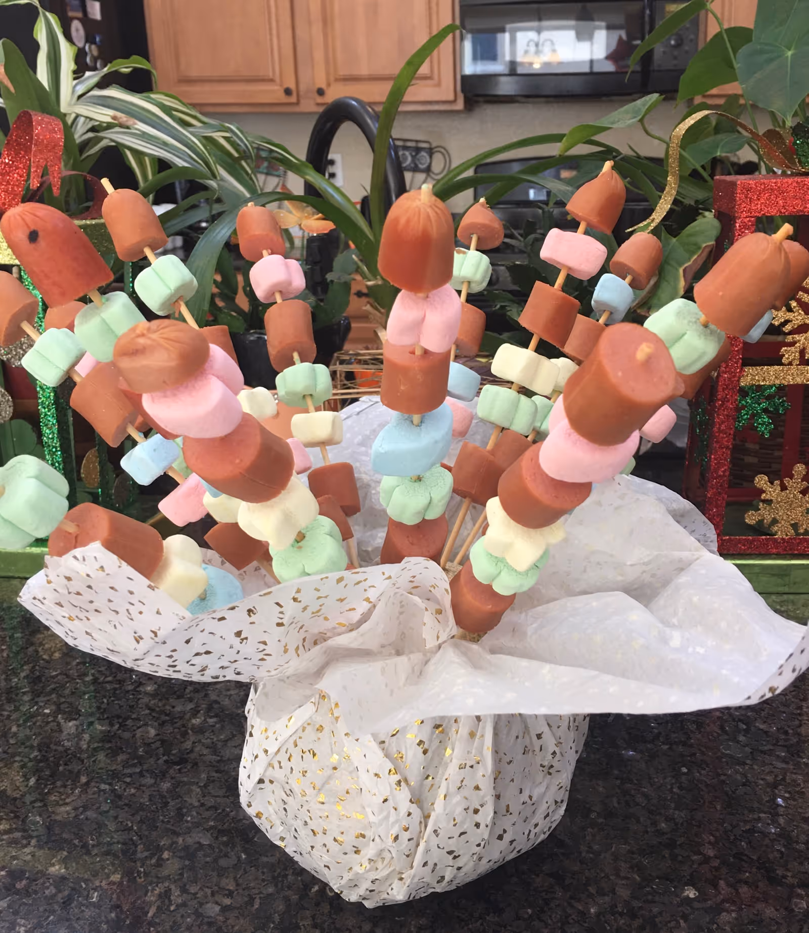 A decorative arrangement of skewers with pieces of hot dogs and colorful marshmallows placed in a white and gold speckled wrapping on a kitchen countertop. In the background, there are green plants and wooden kitchen cabinets.