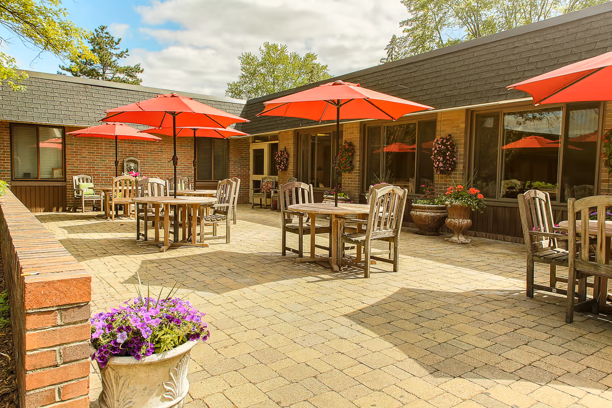 Outdoor patio area with several wooden tables and chairs, each shaded by large red umbrellas. The patio is paved with stone tiles and surrounded by a brick wall and a building with large windows. There are flower pots with purple and red flowers placed around the patio.