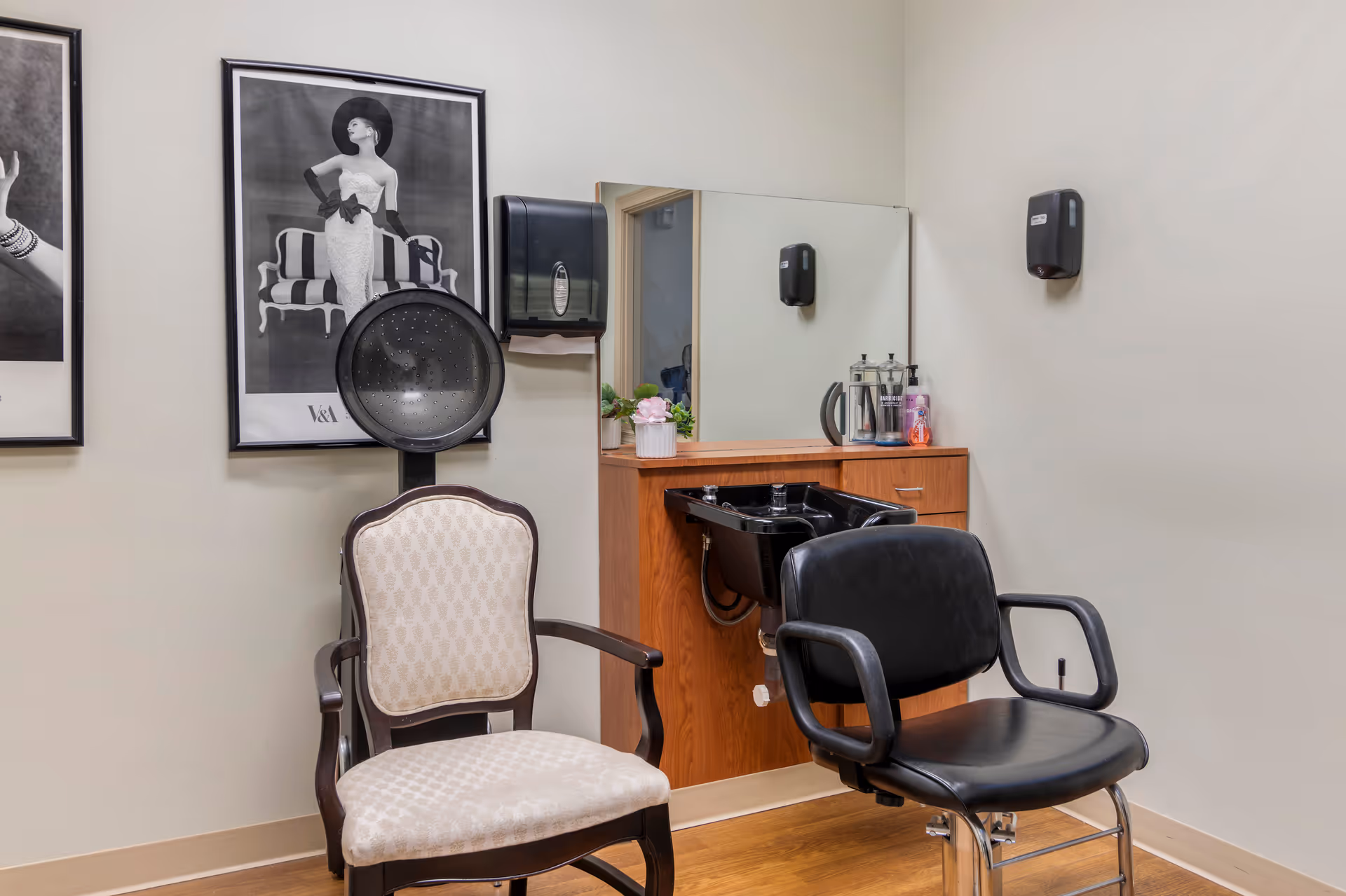 Interior of a hair salon area with a black salon chair and a vintage-style beige upholstered chair. There is a black hair washing sink attached to a wooden cabinet with a large mirror above it. On the wall, there are two black and white framed fashion photographs and two black wall-mounted dispensers.